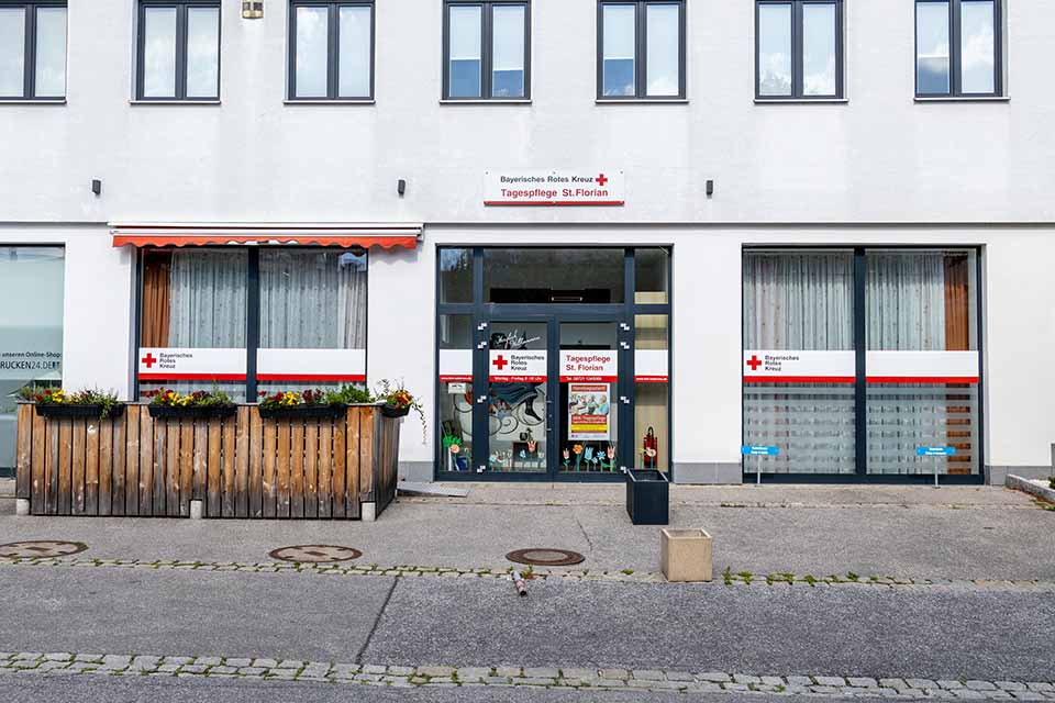 Außenansicht der BRK-Tagespflege St. Florian in Eggenfelden. In der Mitte der Eingang, links davon die Sommerterrasse mit Holzzaun und Blumenkästen mit gelben und roten Blumen. Über der Eingangstür steht groß Bayerisches Rotes Kreuz Tagespflege St. Florian.