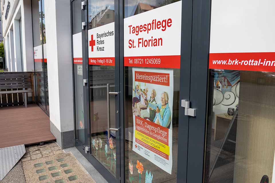Eingangsbereich der BRK-Tagespflege St. Florian in Eggenfelden. Die Türen und Fenster sind im weiß-roten Design des Roten Kreuzes beklebt. Auf der Tür steht in roten Buchstaben Tagespflege St. Florian. Darunter hängt ein Plakat mit dem Titel "hereinspaziert", welches zum kostenlosen Schnuppertag einlädt.