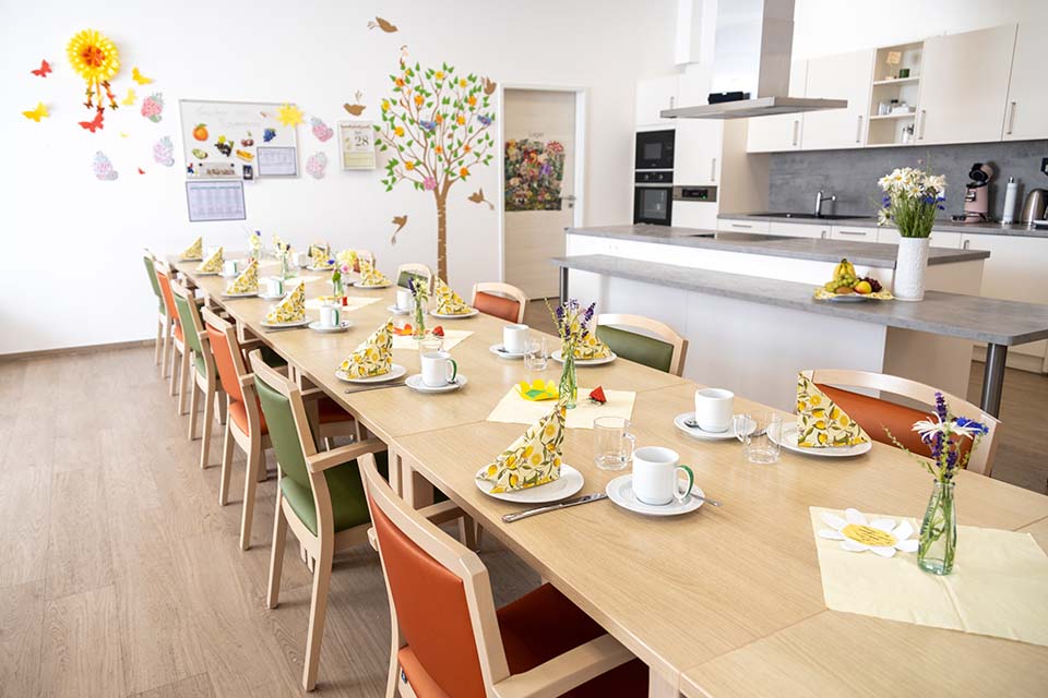 Im großen Gemeinschaftsraum der BRK-Tagespflege St. Florian befindet sich eine lange Tischreihe mit abwechselnd grün und orange bezogenen Holzstühlen die auf beiden Seiten der Tische aufgereiht sind. Die Tische sind für den Nachmittagskaffee dekoriert. Auf den weißen Tellern sind frühlinghafte Servietten zur Dekoration. Die weißen Tassen haben grüne Henkel. Auf der Wand im Hintergrund sind sommerliche Dekorationen zu sehen: Schmetterlinge, die Sonne, Vögel und ein Baum ist an die Wand gemalt. Rechts von dem großen Tisch befindet sich die offene Küche. 