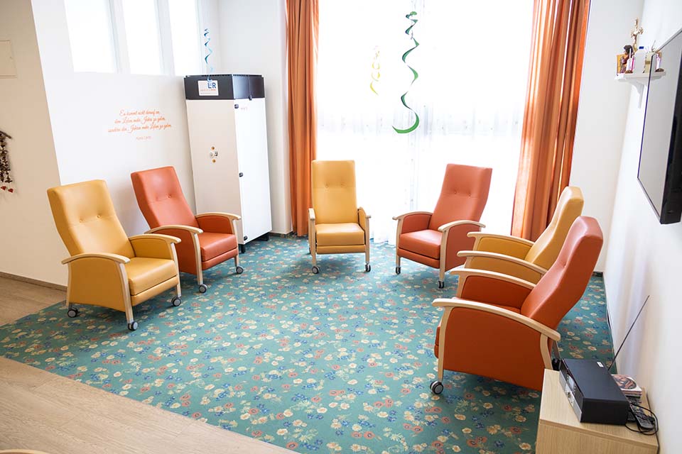 Ein Stuhlkreis aus bequemen Sesseln. & Sessel sind im 3/4 Kreis aufgestellt. Die Sessel sind abwechselnd gelb und orange bezogen. Der Teppich auf dem die Stühle stehen ist grün mit gelben, roten und blauen Blumen. Links im Hintergrund steht eine Lüftungsanalge.
