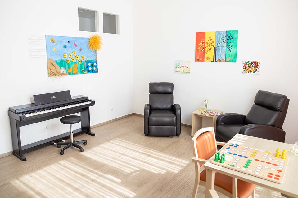 Im Therapieraum der BRK-Tagespflege St. Florian steht an der linken Wand ein Klavier. Darüber hängt ein buntes Bild mit Sonnenblumen. An der Wand daneben hängen drei bunte, sommerliche Bilder. Davor stehen zwei bequeme Ledersessel und dazwischen ein Beistelltisch mit Blumenvase in der Margeriten sind. Im rechten Vordergrund ist ein Tisch mit einem Mensch ärgere dich nicht spiel zu sehen.
