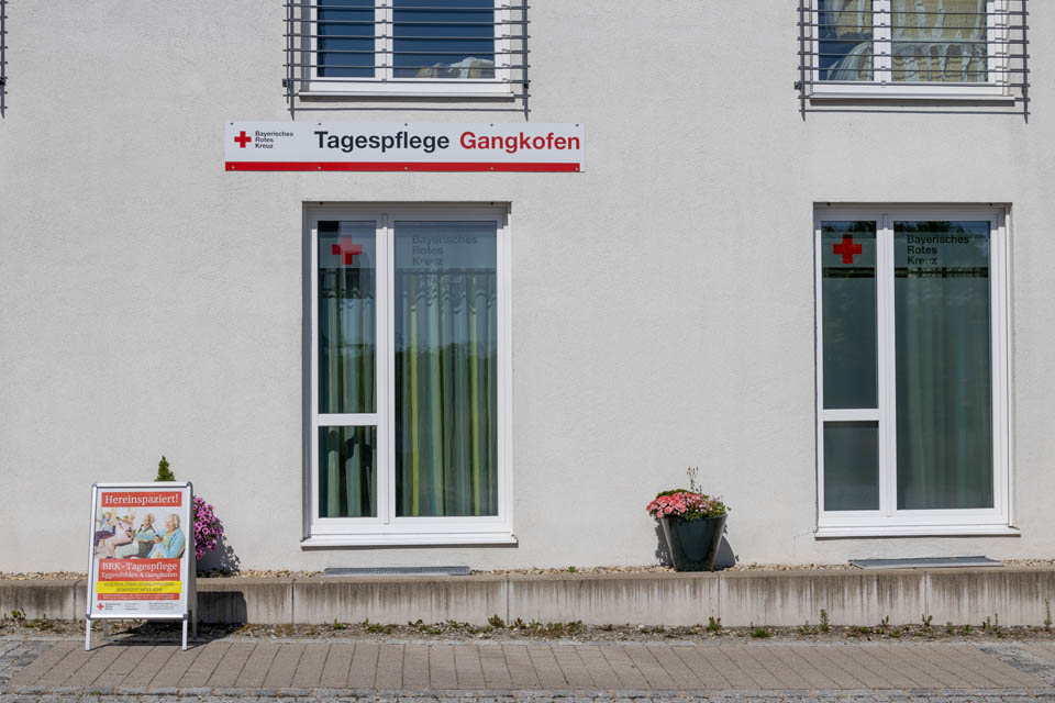 Zwei der hohen Fenster der BRK-Tagespflege in Gangkofen. Die Fenster sind oben mit dem Logo des Bayerischen Roten Kreuzes beklebt. Oberhalb des linken Fensters ist ein breites Schild mit dem BRK-Logo und dem Schriftzug Tagespflege Eggenfelden. links auf dem Gehweg neben dem Fenster steht ein Schild mit der Aufschrift Hereinspaziert um auf den kostenlosen Schnuppertag aufmerksam zu machen.