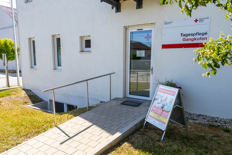 Die Eingangstür zur BRK-Tagespflege in Gangkofen ist seitlich am Gebäude. Die gläserne Türe ist oben mit dem Logo des Bayerischen Roten Kreuzes beklebt. Rechts neben der Tür ist ein großes Schild mit BRK-Logo und dem Schriftzug Tagespflege Gangkofen. Neben dem Gehweg steht ein Schild mit der Aufschrift Hereinspaziert um auf den kostenlosen Schnuppertag aufmerksam zu machen.