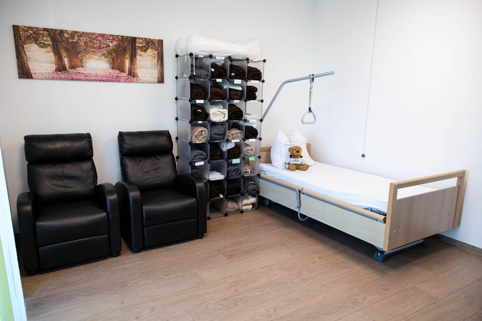 Im Ruheraum der BRK-Tagespflege in Gangkofen befindet sich ein Patientenbett welches verstellbar ist und einen Haltegriff für die Gäste hat. Auf dem Bett sitzt ein Teddybär. Links vom Bett steht ein offenes Regal mit verschiedenen Decken. Rechts vom Regal stehen zwei bequeme Ledersessel. Über den Sesseln ist ein breites Bild mit rosa blühenden Bäumen