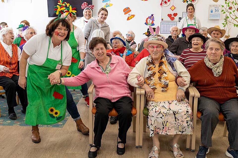 Auf der Faschingsfeier der BRK-Tagespflege Eggenfelden schunkeln die Besucher lachend auf Ihren Plätzen. Einige von Ihnen sind mit Partyhüten oder Perücken verkleidet. Eine Mitarbeiterin der Tagespflege gibt einer Besucherin die Hand und schunkelt mit Ihr. Die Mitarbeiterin trägt eine grüne Schürze die mit Blumen bestickt ist, außerdem hat sie eine Blume ins Gesicht geschminkt und hat trägt einen Blumenhaarreif.
