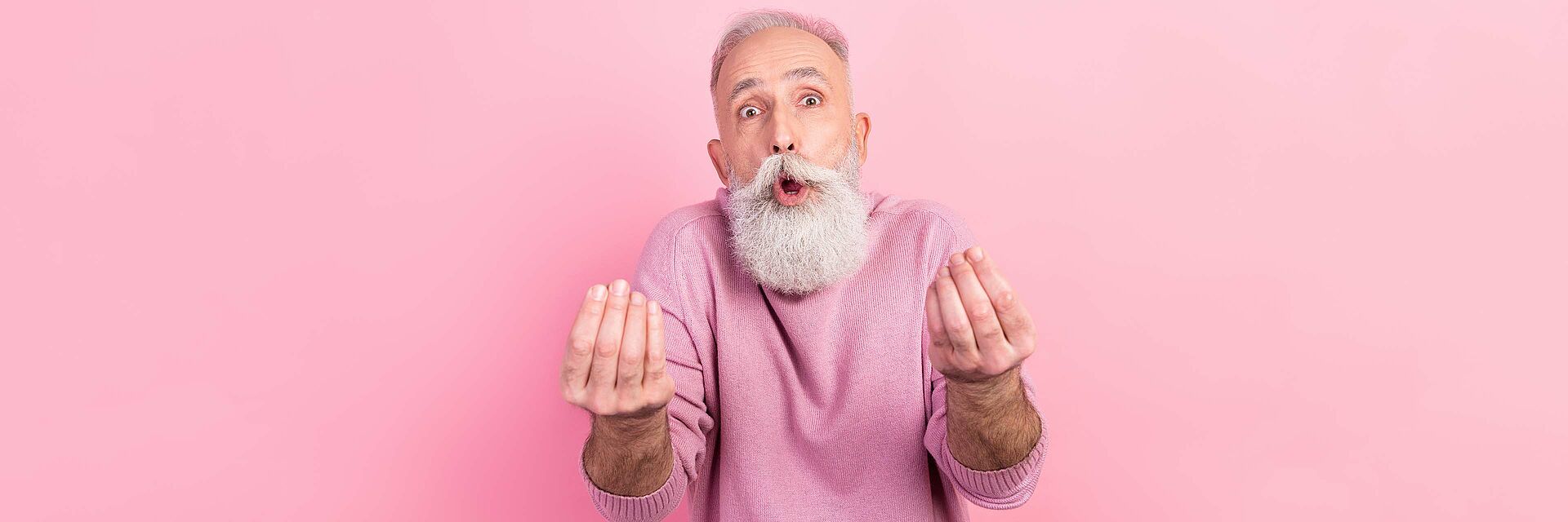 Ein älterer Herr mit weißem langem Bart macht mit seinen Händen das Geld Symbol um die Fragen nach den Kosten für den Besuch der BRK-Tagespflegen Rottal-Inn zu symbolisieren. Der Mann trägt einen rosa Pulli und ist vor einem rosa Hintergrund fotografiert.