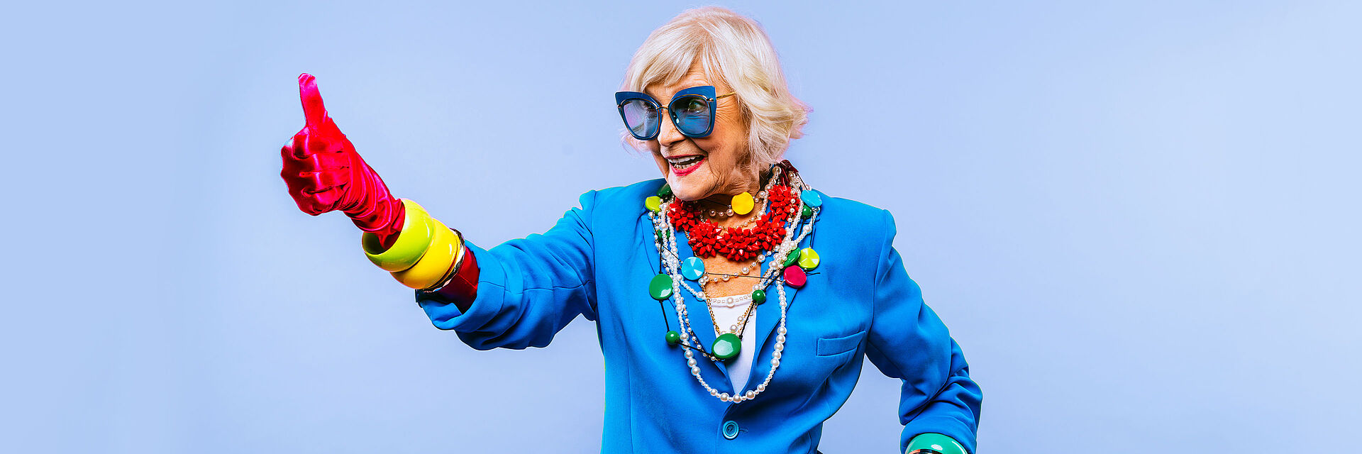 Eine flippige ältere Dame im blauen Sakko und roten Samthandschuhen zeigt den Daumen nach oben und lacht. Sie hat graue Haare und trägt bunte Armreifen und Ketten und dazu eine große Sonnenbrille. 