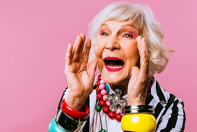 Eine ältere Dame mit weißem Haar nimmt die Hände zum offenen Mund um freudig etwas anzukündigen. Die Frau ist grell geschminkt und trägt bunte große Perlenketten und Armreifen.